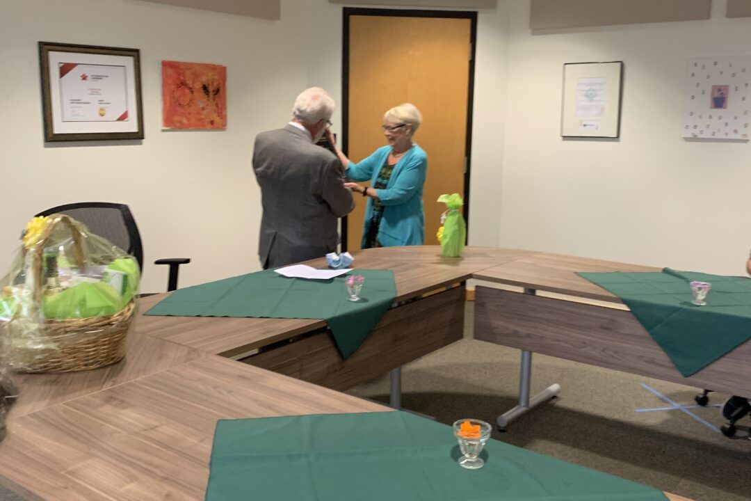 Two individuals shaking hands in a conference room with a basket and face masks on the table.