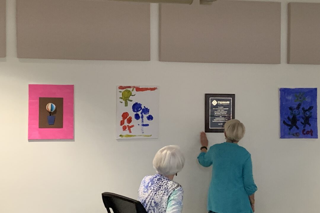 Two individuals observing artwork and a plaque on a gallery wall.