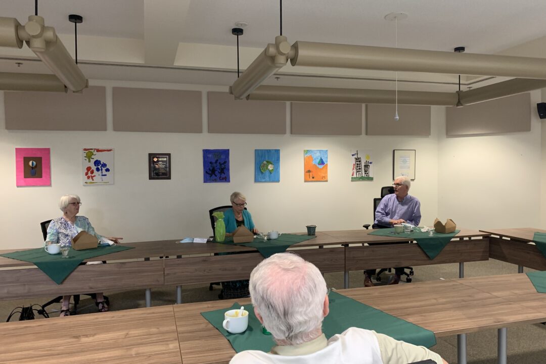 Senior adults attending a meeting at a table with colorful artwork on the wall in the background.