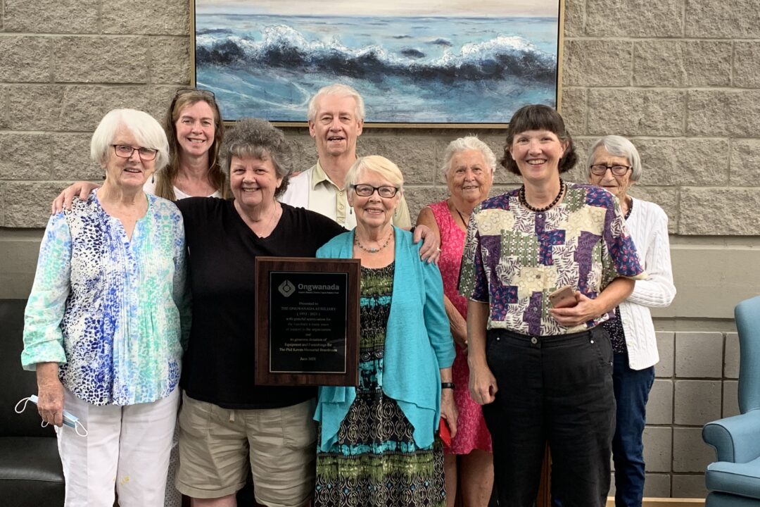 A group of eight smiling adults posing with a commemorative plaque in a room with a large ocean wave painting in the background.