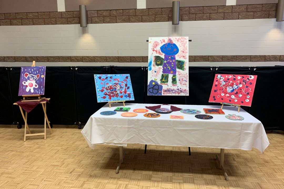 Artwork display at an event featuring colorful paintings and crafted plates arranged on a table and easels.