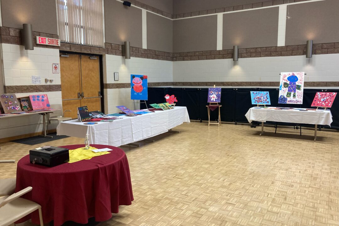 Art exhibition setup in a hall with artwork displayed on easels and tables covered with cloths.