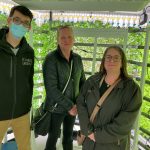 Three people standing in front of a vertical indoor garden with hydroponic plants.