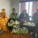 Four individuals posing with trays of packaged greens, possibly during a food distribution event.