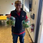 A smiling man wearing glasses, a baseball cap, and a quilted vest, holding two bags of leafy greens indoors.