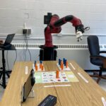 A robotic arm positioned at a table with various blocks and sheets of instructions, presumably used for demonstration or experimentation in a robotics lab.