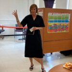 A woman gesturing with her hand in a classroom setting with snacks on a table nearby.