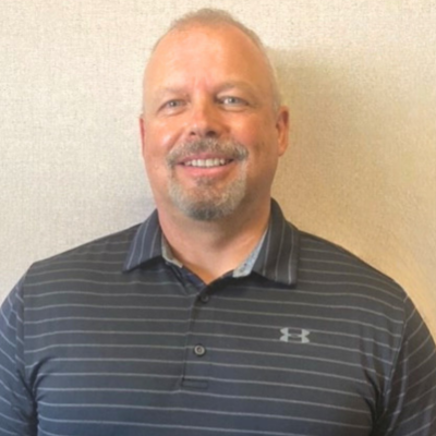 A middle-aged man with a beard smiling in front of a plain wall, wearing a striped polo shirt.