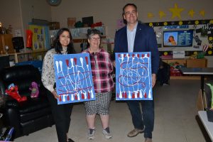 Three adults holding artwork in a classroom decorated with children's educational materials.