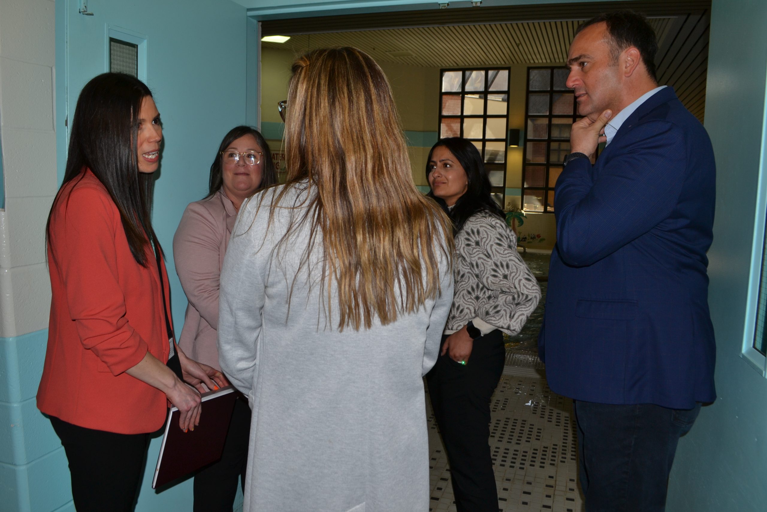 Five adults, three women and two men, engaged in a discussion in a hallway with light blue walls.