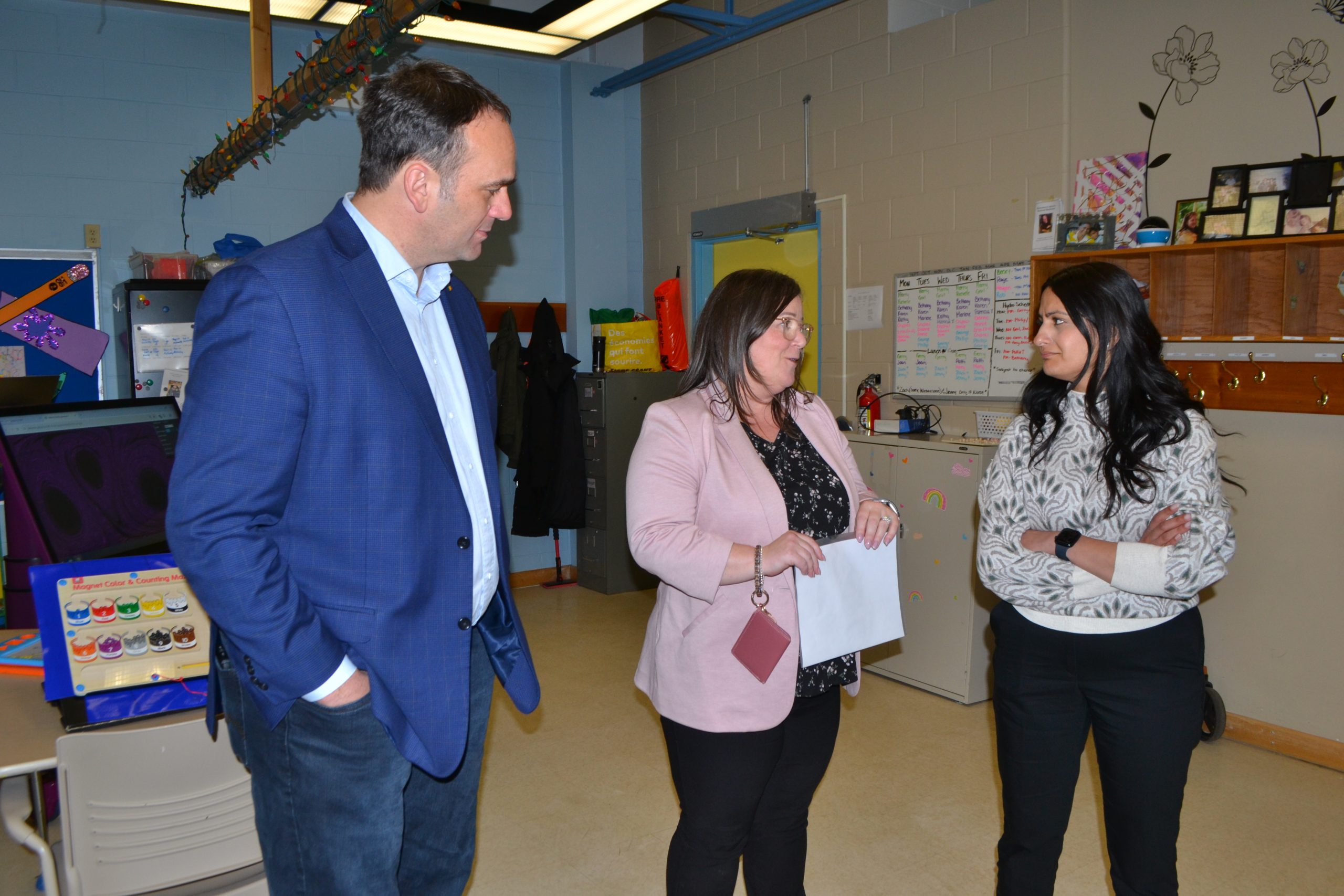 Three adults, two women and one man in business attire, engaged in conversation in a colorful classroom setting.