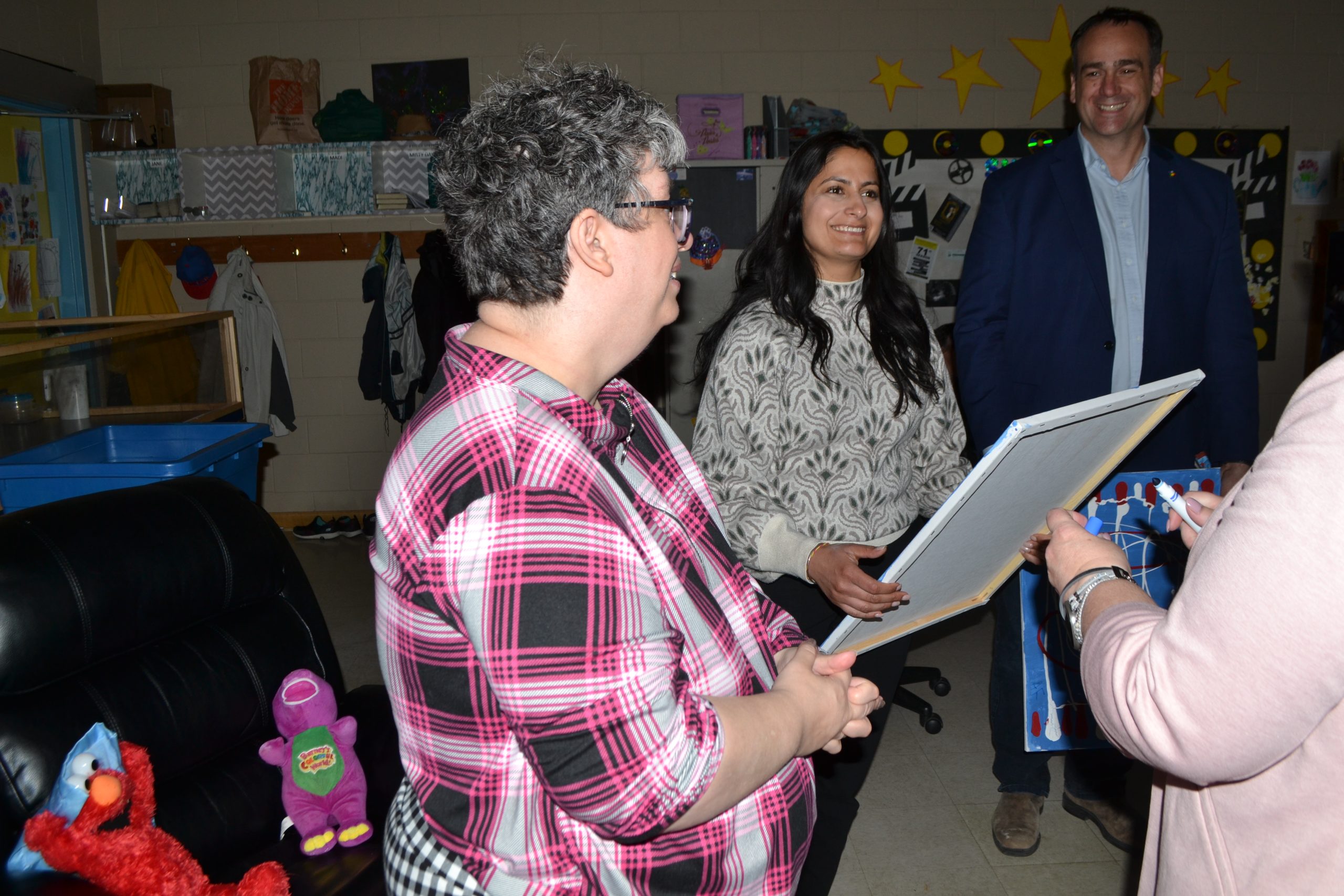 Three adults in a classroom; one woman reading from a clipboard to a man and a woman, with children's toys and artwork in the background.